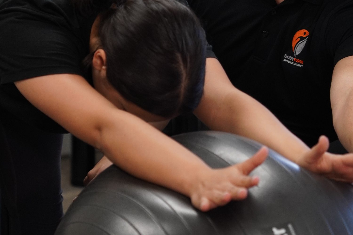 A patient articulating the spine forward over a stability ball, arms reaching long, guided by hand.